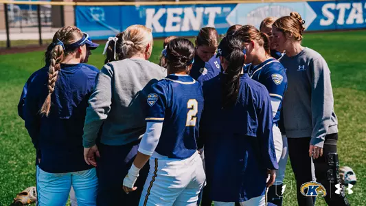 Kent State University softball players and coaching staff gather in a tight huddle on the grass field, viewed from behind. Players are wearing navy blue Kent State MAC conference jerseys and white pants with gold accents, while coaches wear gray Under Armour sweatshirts. Player #2 is visible in the center of the huddle. A Kent State branded banner is visible along the outfield wall in the background. The Ohio outline, Kent State Golden Flashes, and Under Armour logos appear in the bottom right corner.