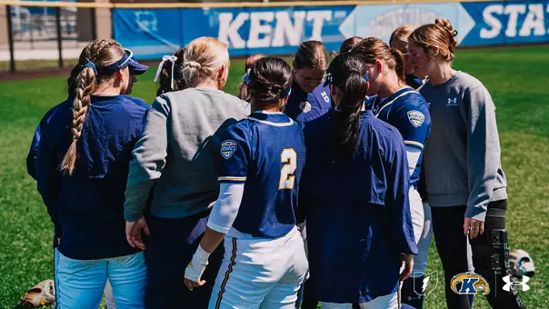 Kent State University softball players and coaching staff gather in a tight huddle on the grass field, viewed from behind. Players are wearing navy blue Kent State MAC conference jerseys and white pants with gold accents, while coaches wear gray Under Armour sweatshirts. Player #2 is visible in the center of the huddle. A Kent State branded banner is visible along the outfield wall in the background. The Ohio outline, Kent State Golden Flashes, and Under Armour logos appear in the bottom right corner.