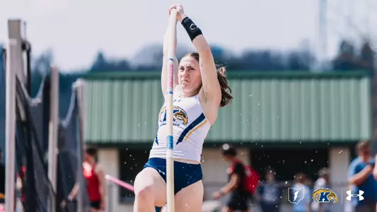 Pole vaulter Ali Krischer grips the pole with both hands raised overhead at the moment of plant, her face showing intense concentration, competing outdoors in a white Kent State uniform with navy shorts. The Kent State Flash logo, Ohio outline, and Under Armour logo appear in the lower right corner.