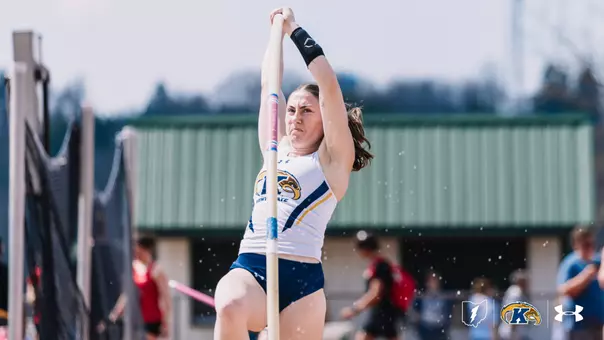 Pole vaulter Ali Krischer grips the pole with both hands raised overhead at the moment of plant, her face showing intense concentration, competing outdoors in a white Kent State uniform with navy shorts. The Kent State Flash logo, Ohio outline, and Under Armour logo appear in the lower right corner.