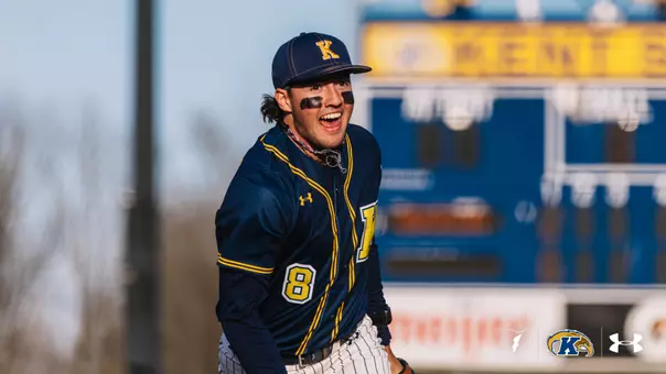 A baseball player wearing a navy blue Kent State uniform with the number 8 visible. They have black eye strips on their cheeks and are standing in front of a scoreboard. The scoreboard displays "Kent" partially. The player appears enthusiastic, and logos from Under Armour are visible at the bottom.