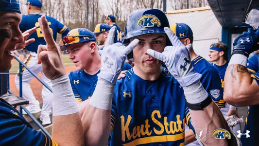 Kent State baseball player in batting helmet and blue-and-gold uniform celebrates in the dugout with teammates after scoring a run, holding up fingers near his face while surrounded by celebrating players