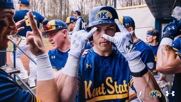 Kent State baseball player in batting helmet and blue-and-gold uniform celebrates in the dugout with teammates after scoring a run, holding up fingers near his face while surrounded by celebrating players