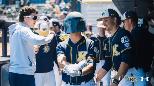 A Kent State Golden Flashes baseball player in a navy 'K' jersey and batting gloves stands in the dugout wearing a Boba Fett helmet as a home run celebration prop, surrounded by laughing teammates in navy Kent State jerseys with MAC patches, and the Kent State lightning bolt, Golden Flashes, and Under Armour logos in the corner.