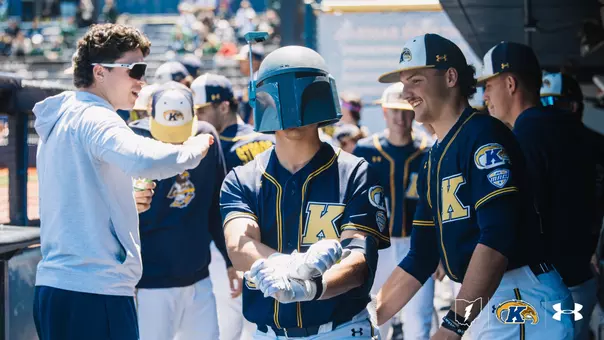 A Kent State Golden Flashes baseball player in a navy 'K' jersey and batting gloves stands in the dugout wearing a Boba Fett helmet as a home run celebration prop, surrounded by laughing teammates in navy Kent State jerseys with MAC patches, and the Kent State lightning bolt, Golden Flashes, and Under Armour logos in the corner.