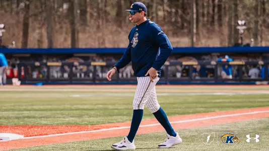 Kent State baseball head coach Jeff Duncan walks along the foul line in a navy Kent State Under Armour pullover, white pinstripe pants, navy socks, a navy gold-brimmed cap, and blue-tinted sunglasses, with the Kent State dugout and baseball wall signage visible in the background.