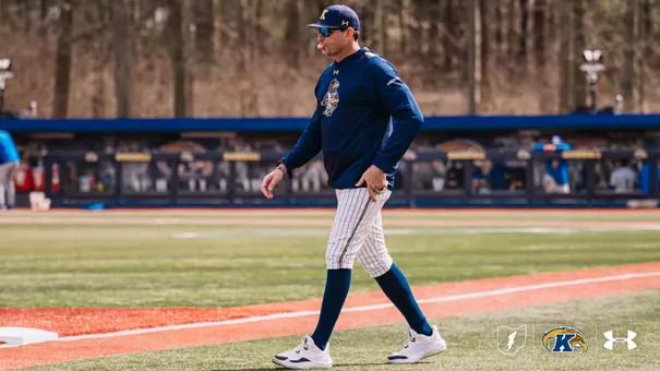 Kent State baseball head coach Jeff Duncan walks along the foul line in a navy Kent State Under Armour pullover, white pinstripe pants, navy socks, a navy gold-brimmed cap, and blue-tinted sunglasses, with the Kent State dugout and baseball wall signage visible in the background.