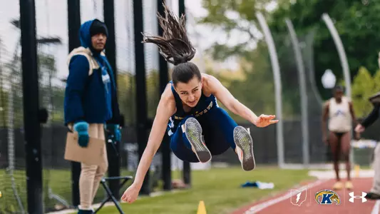 Kent State University track and field student-athlete Monica Naudin executes a long jump, her body nearly parallel to the ground with dark hair streaming upward. She wears a navy blue Kent State Under Armour singlet and navy tights with green-soled spike shoes. A meet official in a blue hoodie watches from the background alongside the red track surface and yellow lane markers. The Kent State Athletics logo and Under Armour wordmark appear in the bottom right corner.