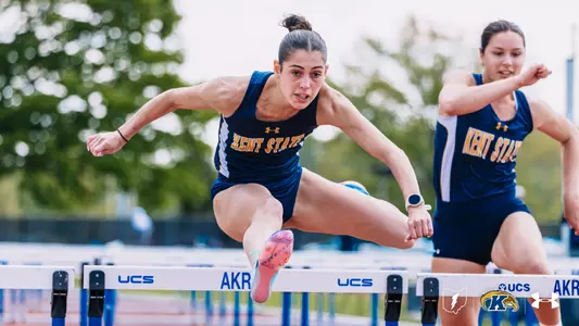 Kent State University track and field student-athlete Alexa Nestor clears a hurdle branded with "UCS" and "AKRON" during a hurdles race. The athlete lunges aggressively over the barrier wearing a navy blue and white Kent State Under Armour singlet, navy briefs, and pink spike shoes; a second Kent State athlete in matching uniform follows closely behind. The Kent State Athletics logo and Under Armour wordmark appear in the bottom right corner.