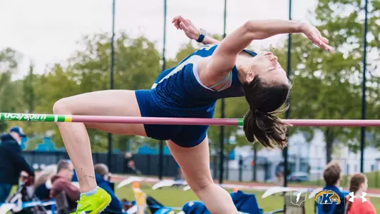 Kent State University track and field student-athlete Matilde Carboncini arches backward over a high jump bar in the Fosbury Flop technique, her back clearing the bar with eyes closed and arms extended. She wears a navy blue and white Kent State Under Armour singlet and navy shorts with neon green spike shoes. A UCS Spirit–branded crossbar is visible. Spectators and trees appear in the blurred background. The Kent State Athletics logo and Under Armour wordmark appear in the bottom right corner.