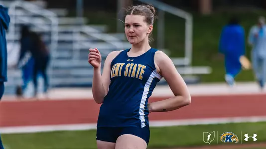 Kent State University track and field student-athlete Ava Koski runs on a red track surface, her hair pulled back in a bun. She wears a navy blue and white Kent State Under Armour singlet and navy shorts. Bleachers are visible in the blurred background. The Kent State Athletics logo and Under Armour wordmark appear in the bottom right corner.