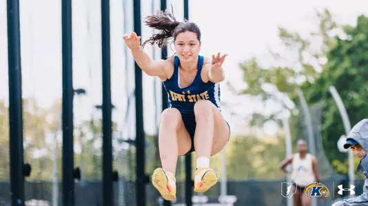 Kent State University track and field student-athlete Matilde Morbin soars through the air during a long jump attempt, arms reaching forward and knees tucked toward her chest. Her dark hair fans out behind her. She wears a navy blue and white Kent State Under Armour singlet and navy shorts with yellow spike shoes. Officials and spectators are visible in the blurred background. The Kent State Athletics logo and Under Armour wordmark appear in the bottom right corner.