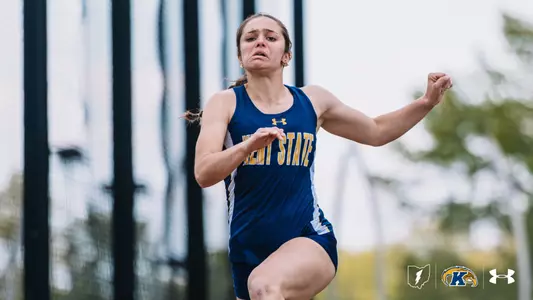 Kent State University track and field student-athlete Maya Justice sprints with intense focus, her right knee driving forward. She wears a navy blue and white Kent State Under Armour singlet and navy shorts. A fenced facility and trees are visible in the blurred background. The Kent State Athletics logo and Under Armour wordmark appear in the bottom right corner.