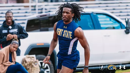 Kent State track and field athlete Dylan Buffum sprints during a race, wearing a navy blue Under Armour sleeveless jersey with "Kent State" in gold lettering and navy blue shorts. His long dreadlocks flow behind him as he drives forward, with spectators and a white vehicle visible in the blurred background. The Kent State Athletics logo and Under Armour wordmark appear in the bottom right corner.