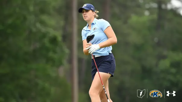 Kent State women's golfer Veronika Kedronová stands on the course in a light blue Kent State polo, navy shorts, and a navy Kent State cap, holding her driver after a tee shot. She gazes down the fairway with a focused expression, with tall green trees blurred in the background. The Ohio state logo, Kent State Golden Flashes logo, and Under Armour logo appear in the bottom right corner.