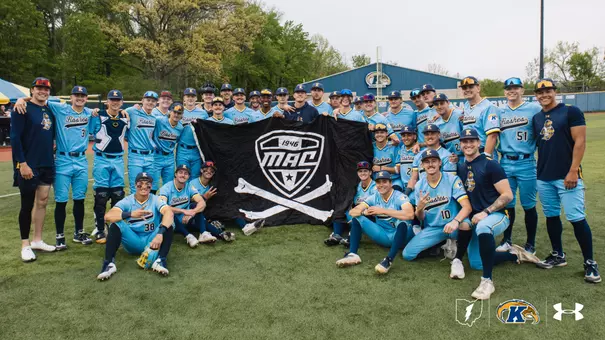 The Kent State baseball team gathers on the field at home for a group photo, holding a large black Mid-American Conference flag with crossed bone, wearing light blue 'Flashes' Under Armour uniforms, with the Kent State baseball facility and championship banner visible on the outfield wall behind them.