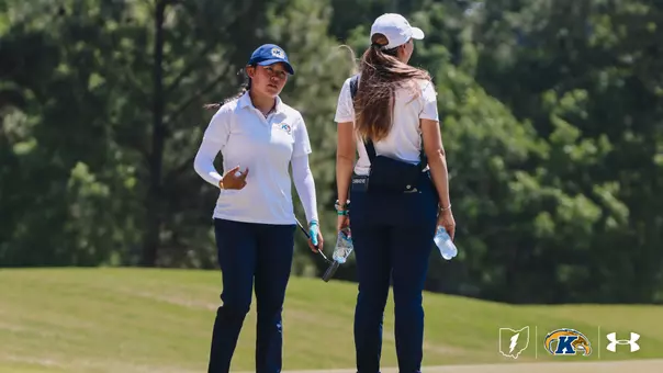 Kent State senior Leon Takagi walks down the fairway in conversation with assistant coach Manuela Carbajo Ré during the final round of the NCAA Chapel Hill Regional at The UNC Finley Golf Course. Takagi, wearing a navy Kent State cap, white polo with a white sun sleeve on her right arm, and navy pants, holds her putter and a water bottle while gesturing with her left hand. Carbajo Ré, seen from behind in a white cap and polo with navy pants and a navy fanny pack labeled "CARBAJO RE," walks alongside her carrying a water bottle. Trees frame the background. The Ohio state logo, Kent State Golden Flashes logo, and Under Armour logo appear in the bottom-right corner.