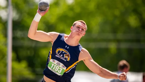 Kent State track and field athlete Linkon Tyrrell, wearing bib no. 1193, winds up to release the shot put during competition. Tyrrell wears a navy blue and gold Kent State sleeveless uniform with an Under Armour logo and a white wrist wrap on his throwing hand. A second athlete is partially visible in the background. Photo features the Kent State Athletics logo and Under Armour wordmark on the uniform.