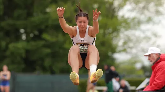 Kent State University track and field athlete Matilde Morrin competes in the long jump at the MAC Outdoor Championships. Wearing bib No. 535, the athlete soars through the air in the hang position with both arms raised and legs tucked forward. Trees and out-of-focus spectators are visible in the background. The Kent State Athletics logo and Under Armour wordmark appear in the lower right corner of the image.