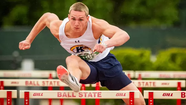 Kent State University track and field Linkon Tyrrell athlete clears a hurdle during competition at Ball State University. The athlete wears a white Under Armour sleeveless uniform top featuring the Kent State Golden Flashes logo and navy blue shorts, with red and white spiked racing shoes. He is captured mid-stride, leaning aggressively forward over the hurdle with an intense expression. Red and white Gill hurdles branded with "Ball State" are visible in the foreground and background, with green foliage blurred in the background. The Kent State Athletics logo and Under Armour wordmark appear on the uniform in the lower portion of the frame.