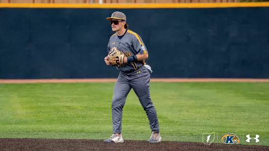 A Kent State Golden Flashes infielder in a gray 'Kent State' Under Armour jersey with gold side stripes, gray pants, sunglasses, and a gray gold-brimmed cap stands in a ready position holding his Wilson glove, with the navy outfield wall and yellow foul line visible behind him.