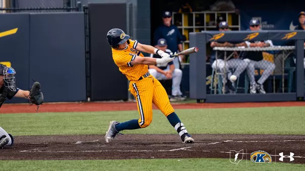 A Kent State Golden Flashes batter makes contact at the plate in a gold pinstripe Under Armour jersey and pants with navy socks and a navy Golden Flashes batting helmet, with the ball visible just off the bat, an opposing catcher at left, and the Toledo dugout with coaches and players watching in the background.