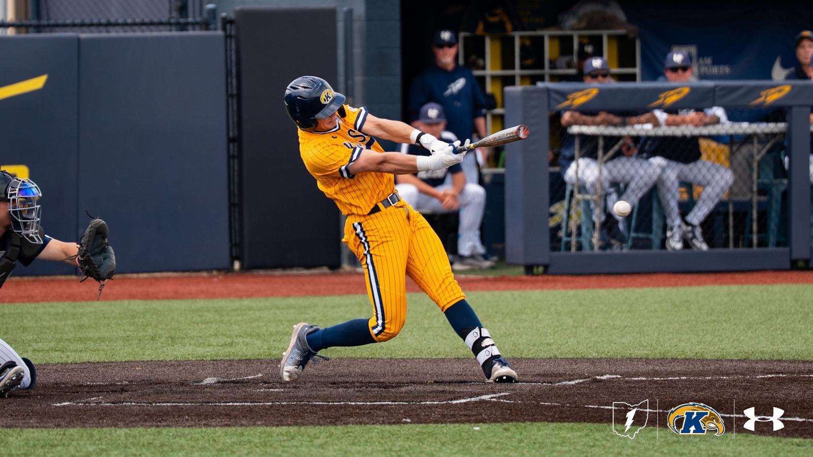 A Kent State Golden Flashes batter makes contact at the plate in a gold pinstripe Under Armour jersey and pants with navy socks and a navy Golden Flashes batting helmet, with the ball visible just off the bat, an opposing catcher at left, and the Toledo dugout with coaches and players watching in th