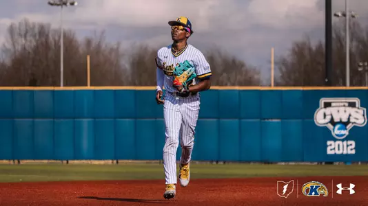 A Kent State Golden Flashes outfielder in a white pinstripe 'Kent State Golden Flashes' Under Armour jersey, white pants, gold cleats, and a navy gold-brimmed cap jogs in from the outfield holding a teal Wilson glove, with the Kent State outfield wall and a 2012 NCAA College World Series banner visible behind him.