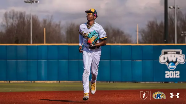 A Kent State Golden Flashes outfielder in a white pinstripe 'Kent State Golden Flashes' Under Armour jersey, white pants, gold cleats, and a navy gold-brimmed cap jogs in from the outfield holding a teal Wilson glove, with the Kent State outfield wall and a 2012 NCAA College World Series banner visible behind him.