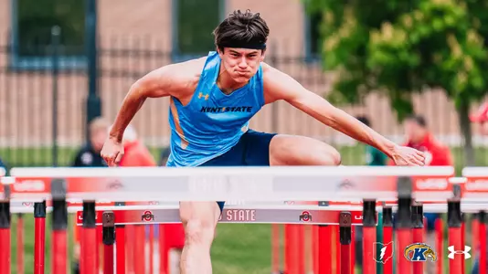 Kent State track and field athlete Beau Harkelroad clears a hurdle during competition at Ohio State, leaning forward with arms extended in mid-stride. He wears a blue Kent State Under Armour singlet and a black headband. Red Ohio State-branded hurdles line the track, with blurred spectators and a brick building visible in the background.