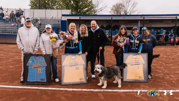Kent State softball Senior Day celebration on the field at Devine Diamond. Three seniors are honored alongside their families. From left, the family of the late Cara Neumann holds a framed light blue number 17 jersey and bouquet of flowers in her memory. In the center, senior Kasey Markusic stands with her family, holding a framed gold number 26 jersey and bouquet of flowers. On the right, senior Brynn Libler stands with her family, holding a framed gold number 3 jersey and bouquet of flowers. A fluffy gray and white dog stands in the center of the group. The dugout and stadium seating with fans are visible in the background. The Ohio Flash, Kent State Golden Flashes, and Under Armour logos appear in the bottom right corner."