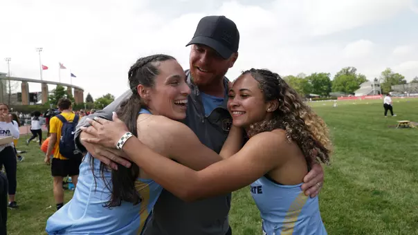 Kent State track and field assistant coach Michael Schober embraces two student-athletes in a group hug on the infield following a competition. A Kent State track and field coach embraces two athletes in a group hug on the infield following a competition. The coach, wearing a dark gray Under Armour quarter-zip and black cap, smiles broadly while hugging the two athletes, who are dressed in light blue and gold Kent State uniforms. Both athletes are smiling and laughing. A track facility with flags, a curved architectural structure, and green trees is visible in the background, with other competitors and spectators milling about.