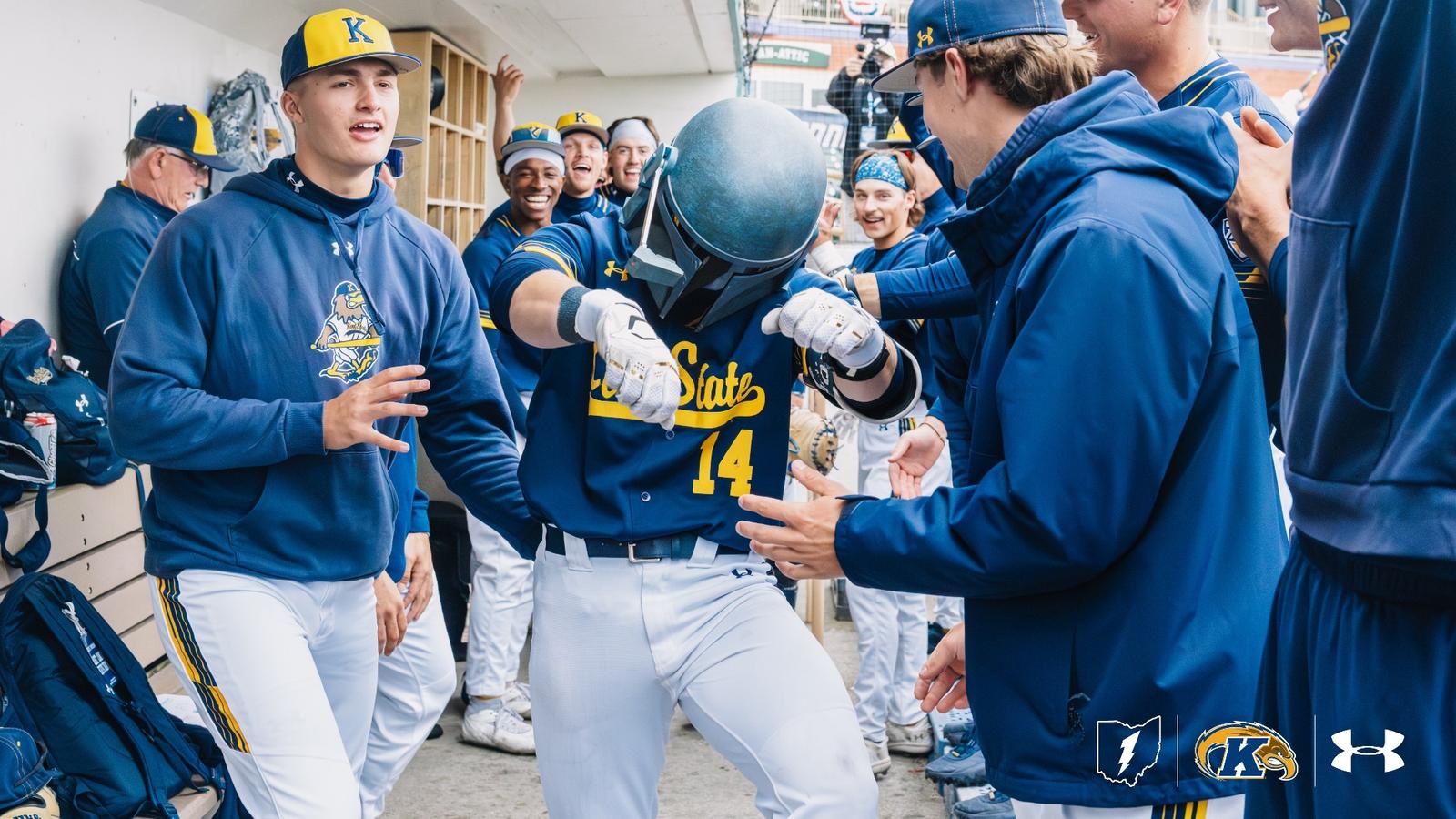Kent State Golden Flashes player wearing No. 14 in a navy 'Kent State' Under Armour jersey is mobbed by jubilant teammates in the dugout while wearing a Boba Fett helmet as a home run celebration prop, with the full dugout erupting in celebration around him and the Kent State lightning bolt, Golden