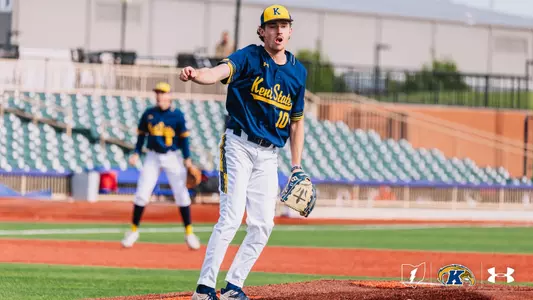 Kent State Golden Flashes pitcher Ciaran Caughey, wearing No. 10 in a navy 'Kent State' Under Armour jersey and white pants, pumps his fist and shouts in celebration on the mound, with a teammate positioned in the infield behind him and a partially filled stadium with teal seats visible in the background.