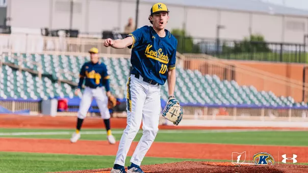 Kent State Golden Flashes pitcher Ciaran Caughey, wearing No. 10 in a navy 'Kent State' Under Armour jersey and white pants, pumps his fist and shouts in celebration on the mound, with a teammate positioned in the infield behind him and a partially filled stadium with teal seats visible in the background.