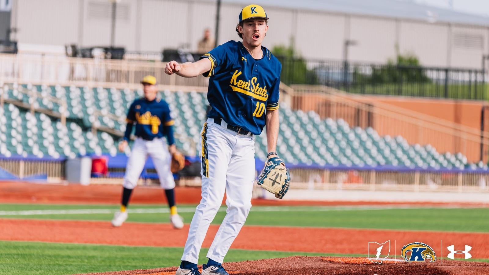 Kent State Golden Flashes pitcher Ciaran Caughey, wearing No. 10 in a navy 'Kent State' Under Armour jersey and white pants, pumps his fist and shouts in celebration on the mound, with a teammate positioned in the infield behind him and a partially filled stadium with teal seats visible in the backg