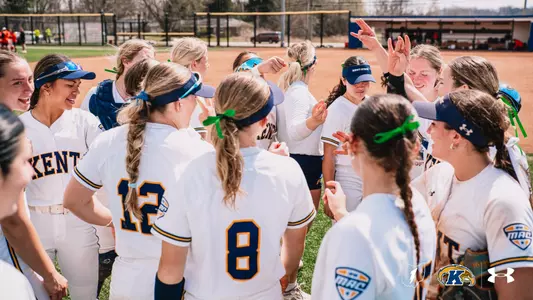 A wide action photo of the Kent State softball team gathered in an energetic team huddle on an outdoor softball field on a sunny day. The players are wearing white Kent State home uniforms with navy and gold lettering, navy and gold visors, and green ribbon hair ties and accessories throughout the group. Several players are visible from behind, including jersey numbers 8 and 12. Players on the right side of the huddle have their hands raised in celebration, with one player making a hand gesture above the group. A catcher's glove is visible on one player at the left. Trees, a red pickup truck, and a dugout structure are visible in the blurred background. The MAC Conference logo appears on the jerseys. The MAC Conference logo, Ohio state outline with lightning bolt, Kent State Golden Flashes logo, and Under Armour logo are displayed in the bottom right corner of the image.