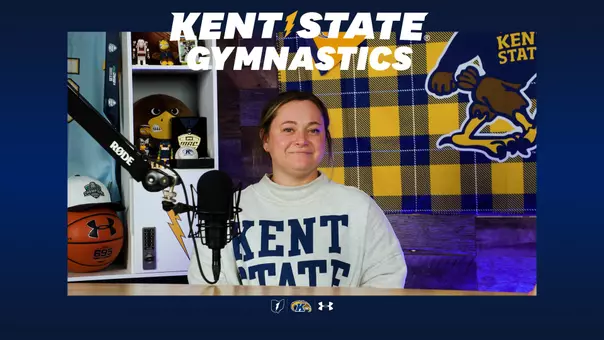Austyn Fobes head gymnastics coach sits at table with microphone for interview. She is wearing a sweatshirt that says Kent State. A 2026 MAC women'ts golf hat is in the background with other kent state giveaways
