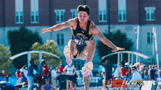 Kent State track and field athlete Luigi Case soars through the air during a long jump attempt, arms spread wide and knees tucked toward his chest. He wears a navy and gold Under Armour uniform with bib number 726 and spiked shoes with pink soles. A crowd of spectators and a brick building are visible in the blurred background. The Kent State Athletics logo and Under Armour wordmark appear in the bottom right corner.