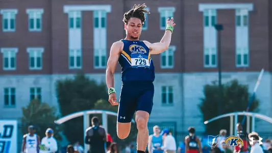 Kent State track and field athlete Amare Hall is captured mid-air during a long jump attempt, his dreadlocks flowing upward as he strides through the jump with one leg extended forward and arms back for balance. He wears a navy and gold Under Armour uniform with bib number 729, navy shorts, black socks, and green wristbands on both wrists. A crowd of spectators and a brick building are visible in the blurred background. The Kent State Athletics logo and Under Armour wordmark appear in the bottom right corner.