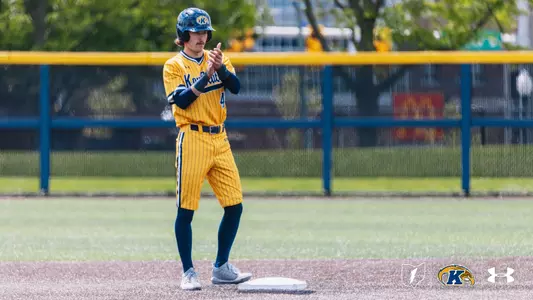 A Kent State Golden Flashes baserunner stands on second base clapping, wearing a gold pinstripe 'Kent State' Under Armour jersey, gold pinstripe pants, navy sleeves and socks, and a navy Golden Flashes batting helmet, with the outfield fence and trees visible in the background.