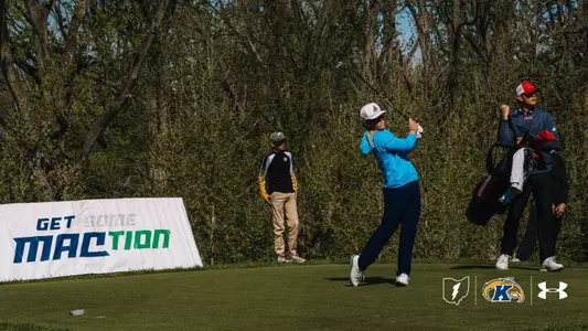 Kent State golfer Liam Curtis follows through on his swing wearing a bright blue pullover and white cap, with his caddie standing to his right and a "Get Some MAC-tion" banner visible behind him on the tee box.