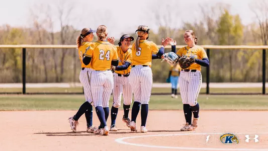 Five Kent State softball players celebrate together on the infield dirt, exchanging high-fives after a play. The players are wearing gold Kent State jerseys with white pinstripe pants and navy socks. Visible jersey numbers include 24 and 6. A fielding glove is visible on one player's hand. A sunny, tree-lined outfield fence is visible in the background with an opposing player in light blue visible in the distance. The Ohio Flash, Kent State Golden Flashes, and Under Armour logos appear in the bottom right corner.