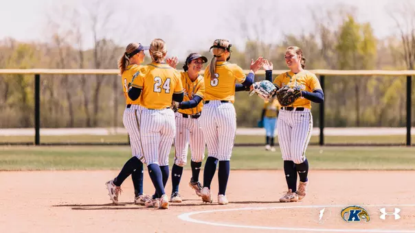 Five Kent State softball players celebrate together on the infield dirt, exchanging high-fives after a play. The players are wearing gold Kent State jerseys with white pinstripe pants and navy socks. Visible jersey numbers include 24 and 6. A fielding glove is visible on one player's hand. A sunny, tree-lined outfield fence is visible in the background with an opposing player in light blue visible in the distance. The Ohio Flash, Kent State Golden Flashes, and Under Armour logos appear in the bottom right corner.
