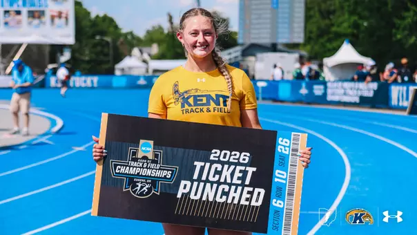 A Kent State track and field athlete smiles while holding a large "2026 Ticket Punched" sign on a blue track at the NCAA DI Outdoor Track & Field Championships regional site. The sign features the 2026 NCAA DI Track & Field Championships logo and notes the meet location of Eugene, Ore. The athlete wears a gold Kent Track & Field t-shirt with the Under Armour logo. A Kentucky banner is visible on the track railing in the background. The Kent State Athletics logo and Under Armour wordmark appear in the bottom right corner of the image.