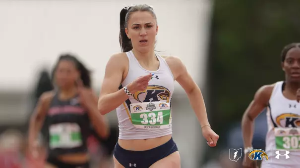 Casey Musgrave of Kent State drives through a sprint event, wearing a white Kent State Under Armour tank top and navy shorts with bib No. 334. A teammate wearing bib No. 303 and a competitor in a dark uniform are visible in the background. The Kent State Athletics logo and Under Armour wordmark appear in the lower right corner.