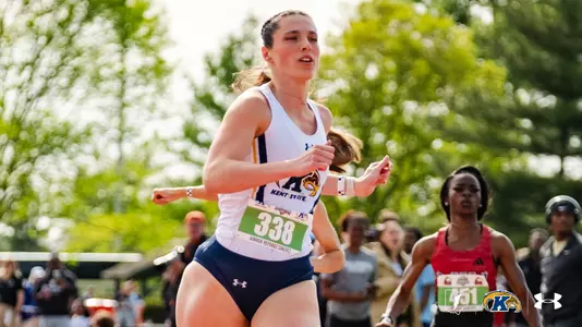 Ainhoa Reparaz Sanchez of Kent State runs in a distance event, wearing a white Kent State Under Armour tank top and navy shorts with bib No. 338. A competitor in a red uniform trails in the background. The Kent State Athletics logo and Under Armour wordmark appear in the lower right corner.