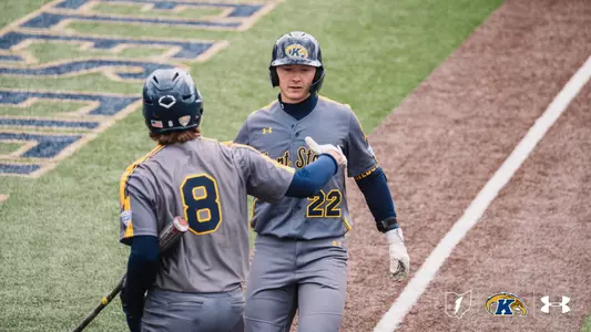 Kent State Golden Flashes player No. 22 exchanges a handshake with teammate No. 8 near home plate after scoring a run, both wearing gray 'Kent State' Under Armour jerseys and gray pants, with the Kent State 'K' logo visible in the outfield grass behind them.
