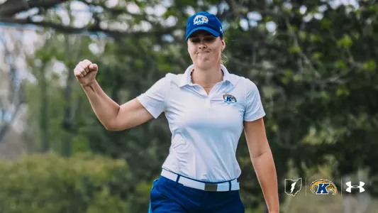 Veronika Kedronová pumps her fist in celebration on the golf course. She wears a white Kent State golf polo, blue Kent State cap and blue pants, with mature trees in soft focus behind her. The Kent State Athletics Ohio mark, primary athletic logo and Under Armour logo are arranged in the bottom-right corner.