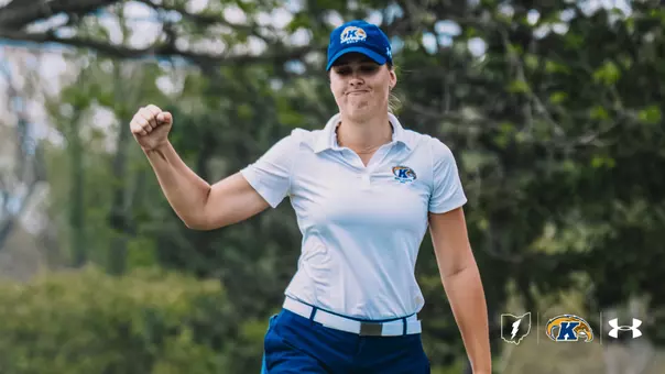 Veronika Kedronová pumps her fist in celebration on the golf course. She wears a white Kent State golf polo, blue Kent State cap and blue pants, with mature trees in soft focus behind her. The Kent State Athletics Ohio mark, primary athletic logo and Under Armour logo are arranged in the bottom-right corner.